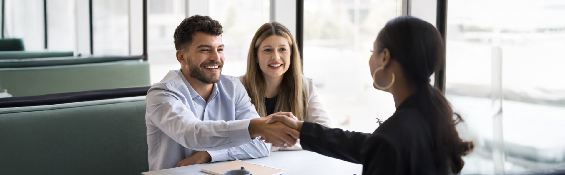 Three people sitting at a table in a meeting. Two are smiling and shaking hands with each other.